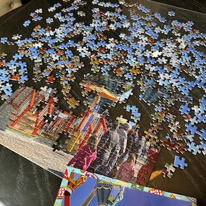 May include: A partially completed jigsaw puzzle featuring a colorful amusement park scene with rides, buildings, and people. The puzzle pieces are scattered on a glass table. The puzzle box is visible in the lower right corner of the image.