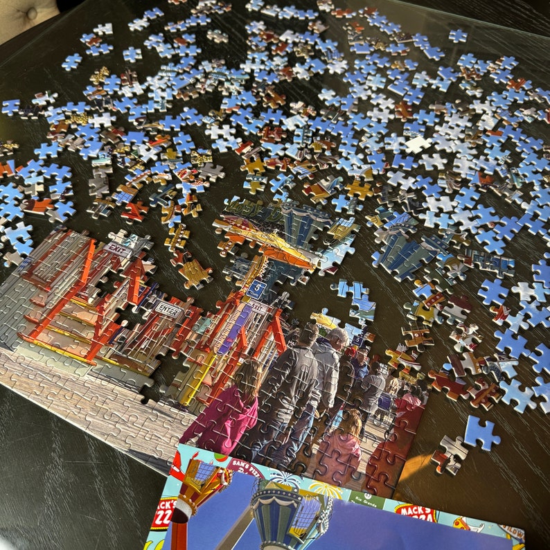May include: A partially completed jigsaw puzzle featuring a colorful amusement park scene with rides, buildings, and people. The puzzle pieces are scattered on a glass table. The puzzle box is visible in the lower right corner of the image.