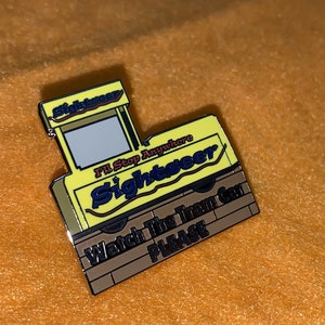 Puede incluir: Un pin de esmalte amarillo y marr&oacute;n con forma de autob&uacute;s tur&iacute;stico. El pin tiene las palabras "Sightseer" e "I'll Stop Anywhere" en una fuente estilizada. La parte inferior del pin dice "Watch The Tram Car PLEASE".
