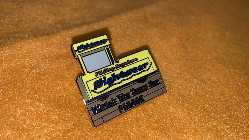 Puede incluir: Un pin de esmalte amarillo y marr&oacute;n con forma de autob&uacute;s tur&iacute;stico. El pin tiene las palabras "Sightseer" e "I'll Stop Anywhere" en una fuente estilizada. La parte inferior del pin dice "Watch The Tram Car PLEASE".