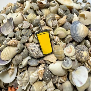 K&ouml;nnte beinhalten: Eine gelbe Emaille-Anstecknadel in Form einer Tasse mit einem Strohhalm, umgeben von einem Haufen Muscheln.