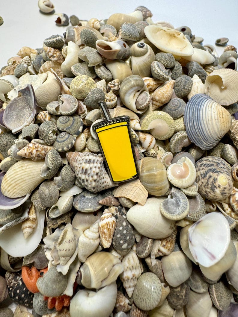 K&ouml;nnte beinhalten: Eine gelbe Emaille-Anstecknadel in Form einer Tasse mit einem Strohhalm, umgeben von einem Haufen Muscheln.