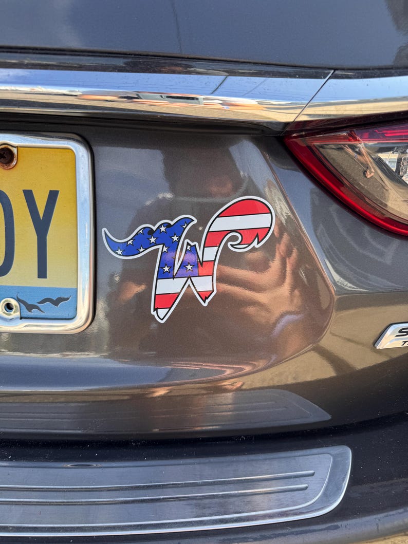 May include: A sticker with a patriotic design, featuring the letter "W" in red and white stripes and blue with white stars. The sticker is affixed to the rear of a gray car.