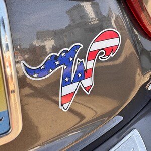 May include: A car sticker featuring a stylized letter "W" with an American flag design. The left side of the "W" is blue with white stars, and the right side has red and white stripes. The sticker is affixed to a gray vehicle.