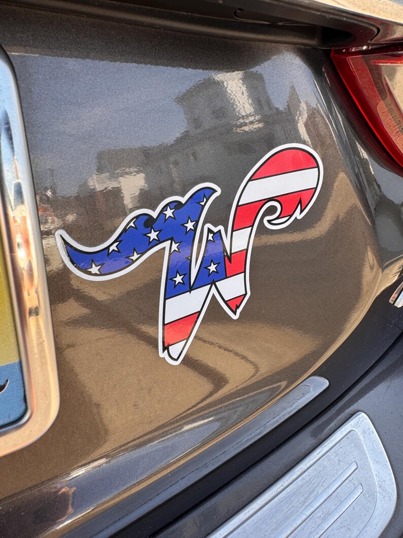 May include: A car sticker featuring a stylized letter "W" with an American flag design. The left side of the "W" is blue with white stars, and the right side has red and white stripes. The sticker is affixed to a gray vehicle.