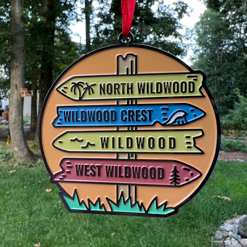 May include: A round, orange, metal ornament with a signpost design. The signpost has four signs pointing in different directions. The signs read "North Wildwood", "Wildwood Crest", "Wildwood", and "West Wildwood".