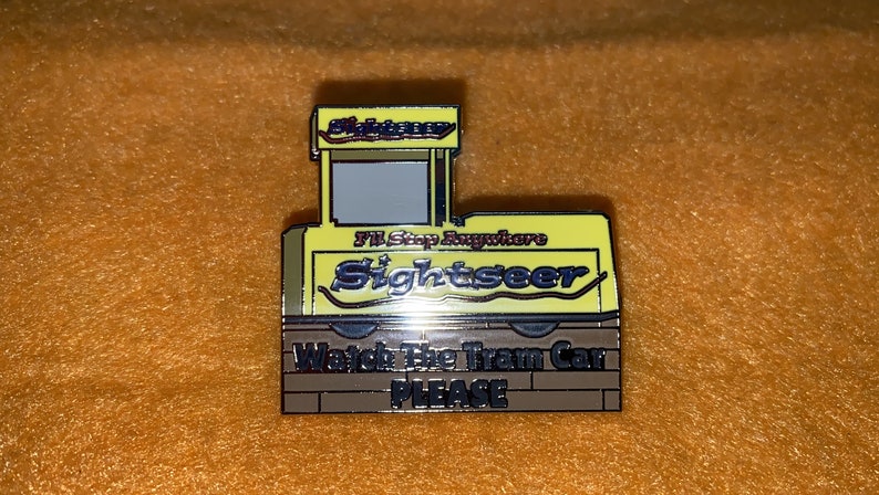 Puede incluir: Un pin de esmalte amarillo y marr&oacute;n con forma de tranv&iacute;a. El pin presenta las palabras "Sightseer" y "I'll Stop Anywhere". La parte inferior del pin dice "Watch The Tram Car PLEASE".