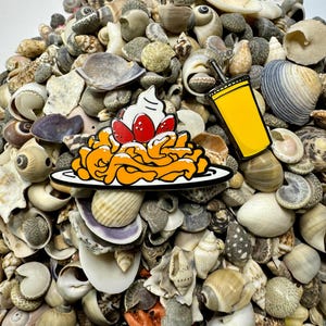 K&ouml;nnte beinhalten: Zwei Emaille-Pins auf einem Bett aus Muscheln. Ein Pin ist ein gelbes Getr&auml;nk mit einem Strohhalm und der andere ist ein Teller mit orangefarbenen und wei&szlig;en Wirbeln mit roten Kirschen und wei&szlig;er Schlagsahne.