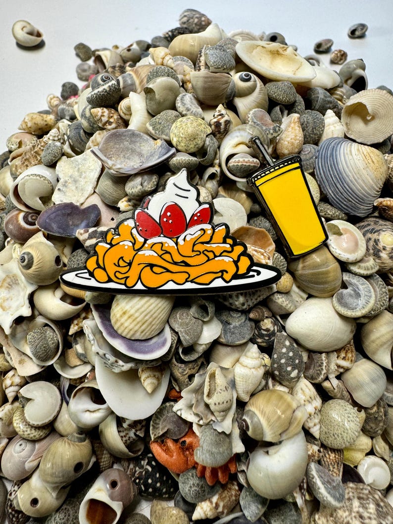 K&ouml;nnte beinhalten: Zwei Emaille-Pins auf einem Bett aus Muscheln. Ein Pin ist ein gelbes Getr&auml;nk mit einem Strohhalm und der andere ist ein Teller mit orangefarbenen und wei&szlig;en Wirbeln mit roten Kirschen und wei&szlig;er Schlagsahne.
