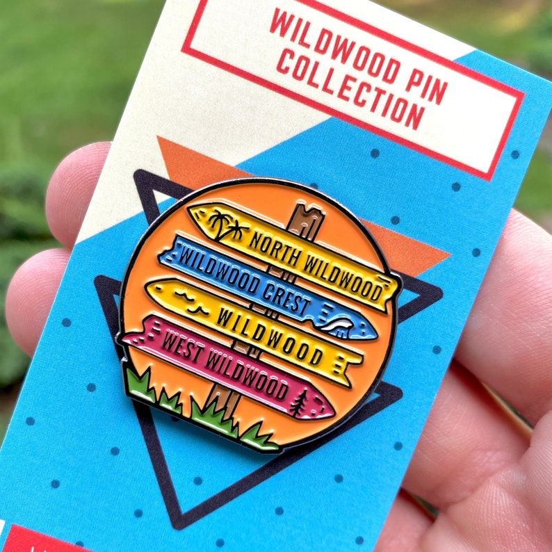 Pu&ograve; includere: Una spilla smaltata colorata con un cartello stradale con quattro frecce direzionali che puntano verso diverse aree di Wildwood, New Jersey. Le frecce indicano "North Wildwood", "Wildwood Crest", "Wildwood" e "West Wildwood".
