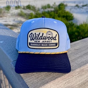 May include: A light blue baseball cap with a dark navy blue brim and a yellow rope detail. The front features a rectangular patch with the text Wildwood New Jersey Boardwalk Beach Summer and a graphic of a Ferris wheel in black and yellow.