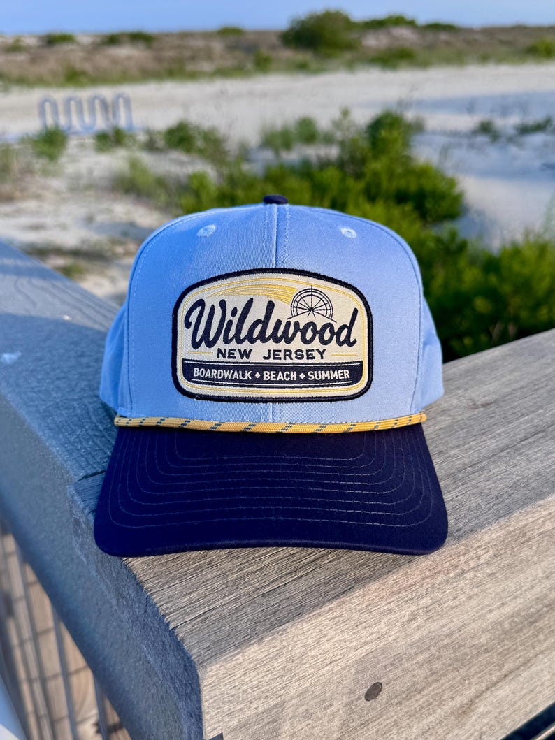 May include: A light blue baseball cap with a dark navy blue brim and a yellow rope detail. The front features a rectangular patch with the text Wildwood New Jersey Boardwalk Beach Summer and a graphic of a Ferris wheel in black and yellow.