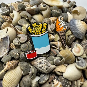 Op de afbeelding: Drie emaille pinnen met een blauwe container met friet, een rode ketchupbeker en een oranje ijshoorn op een bed van schelpen.