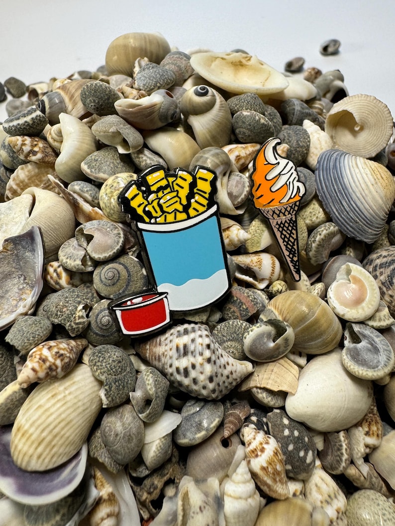 Op de afbeelding: Drie emaille pinnen met een blauwe container met friet, een rode ketchupbeker en een oranje ijshoorn op een bed van schelpen.