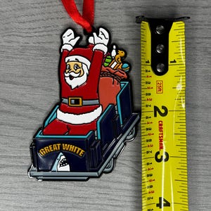 May include: A red and white Santa Claus ornament riding a blue roller coaster with the words "Great White" on the side. The ornament has a red ribbon attached to the top.