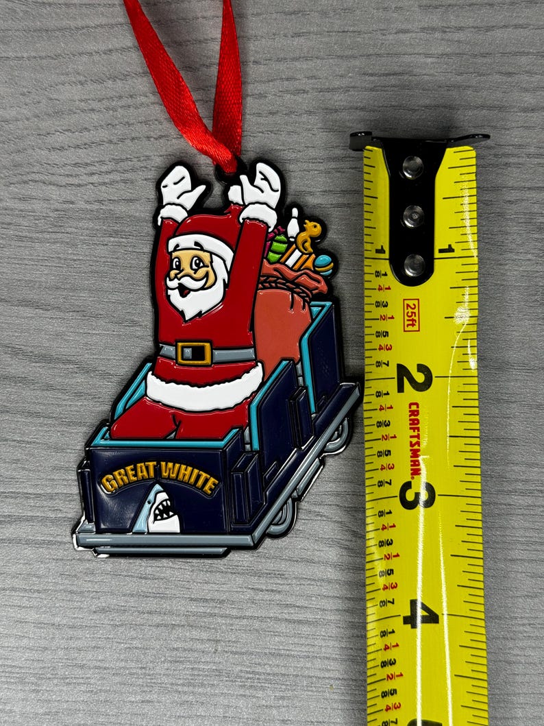 May include: A red and white Santa Claus ornament riding a blue roller coaster with the words "Great White" on the side. The ornament has a red ribbon attached to the top.