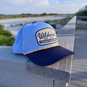 May include: A light blue baseball cap with a dark navy blue brim and a yellow rope detail. The front features a patch with the text Wildwood New Jersey Boardwalk Beach Summer and a small graphic of a Ferris wheel on a cream background.