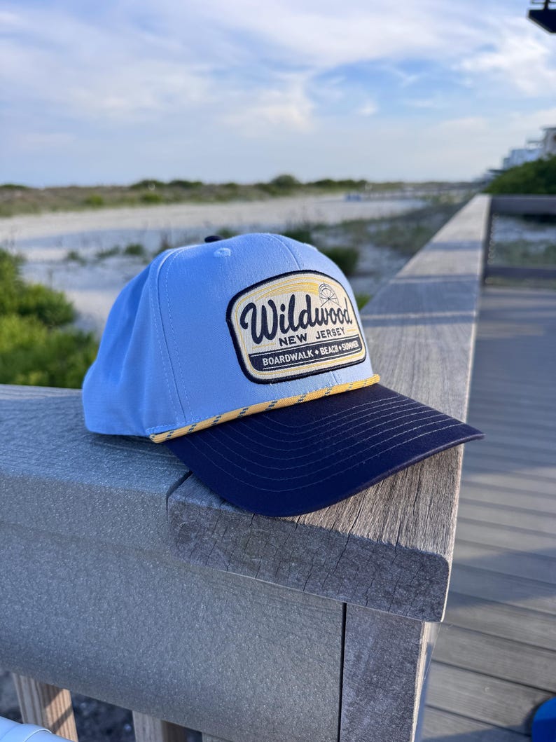 May include: A light blue baseball cap with a dark navy blue brim and a yellow rope detail. The front features a patch with the text Wildwood New Jersey Boardwalk Beach Summer and a small graphic of a Ferris wheel on a cream background.