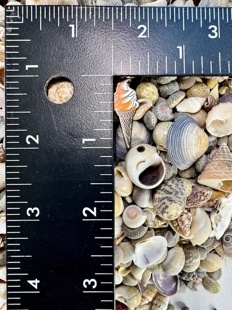 Op de afbeelding: Een close-up van een kleine oranje en witte ijshoorn pin op een bed van schelpen. De pin is ongeveer 2,5 cm hoog en is gemaakt van metaal.