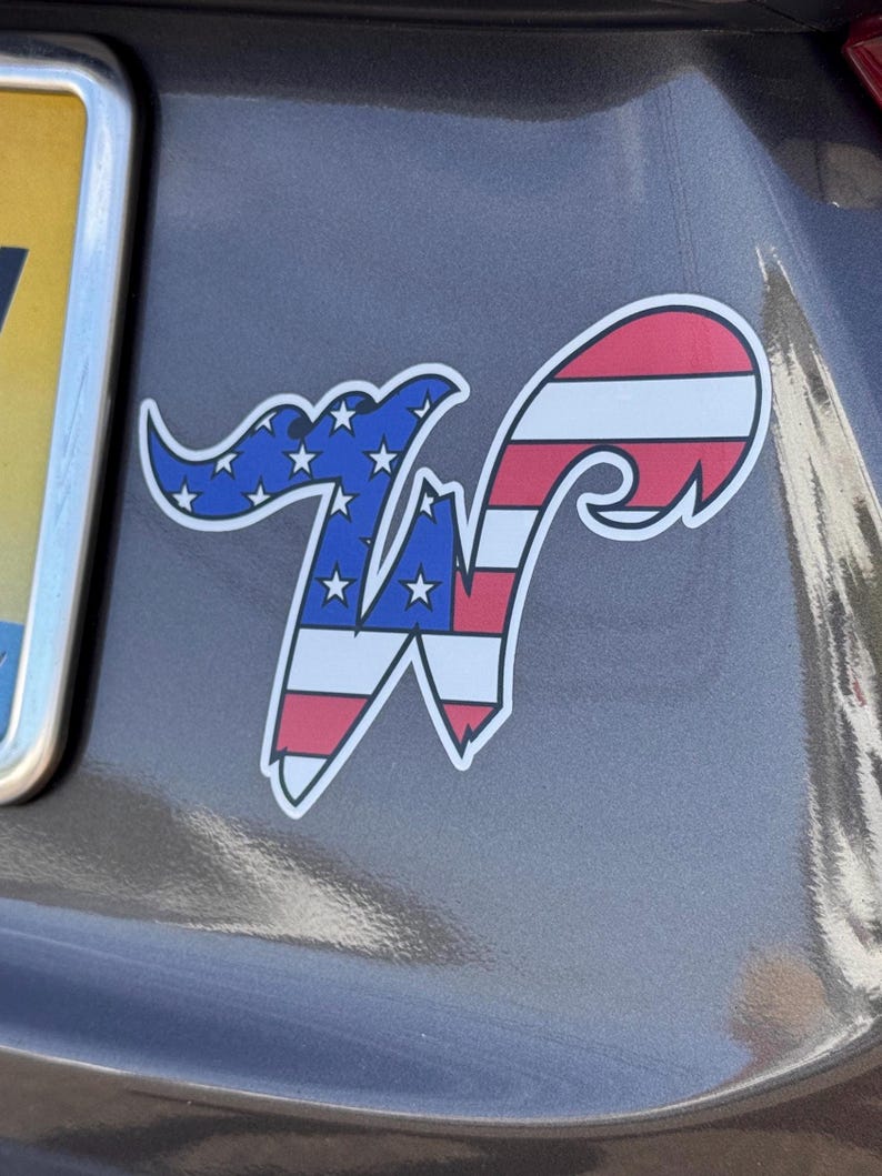 May include: A white-bordered sticker featuring a stylized letter "W" with an American flag design. The top portion is blue with white stars, and the bottom is red and white stripes. The sticker is affixed to a gray surface.