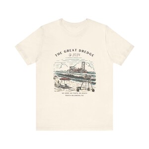 May include: Cream-colored t-shirt featuring a beach scene with the text "THE GREAT DREDGE OF 2024." The graphic includes a lifeguard stand, waves, and a dredging machine. Below the image, it reads "NO SAND, NO TENTS, NO BEACH NORTH WILDWOOD, NJ."