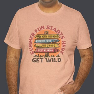 Pu&ograve; includere: Una maglietta color pesca con una grafica circolare che recita "Summer Fun Starts Here". La grafica presenta segnali direzionali che puntano a North Wildwood, Wildwood Crest, Wildwood e West Wildwood. Il testo "Get Wild" &egrave; in basso.