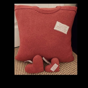 May include: A red knitted cushion with a white label that reads "This is the jumper I used to wear Whenever you hold it Know that I am there". Two smaller red heart-shaped cushions are in front of the larger cushion.