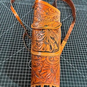 Puede incluir: Un carcaj de cuero marrón con intrincados detalles florales. Tiene una correa para el hombro y un pequeño bolsillo en la parte delantera. El carcaj está diseñado para guardar flechas.