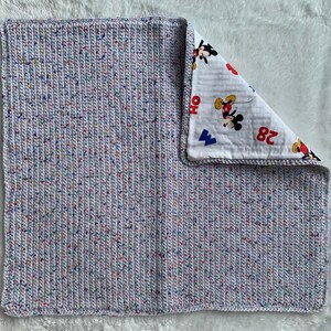 May include: A square baby blanket with a light grey speckled knit outer layer. The inner layer is white fabric printed with a Mickey Mouse pattern in red, black, and yellow. The blanket is soft and appears to be handmade.