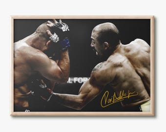 May include: A framed photograph of two mixed martial arts fighters in a match. One fighter is throwing a punch while the other defends. The image features a gold-colored signature on the lower right side and is encased in a light wood frame.