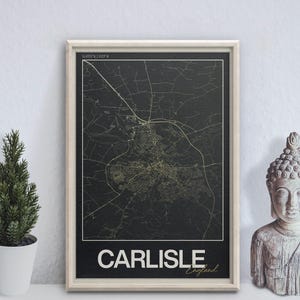 May include: A framed map of Carlisle, England, in black and gold. The city map is displayed with a white border and the city name at the bottom. A small decorative tree and a Buddha statue are on either side.