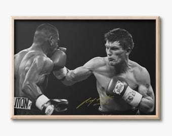 May include: A black and white framed photograph of a boxing match featuring two athletes in motion. One athlete is throwing a punch while wearing boxing gloves with the Lonsdale logo. A gold signature is visible on the lower right side of the print.