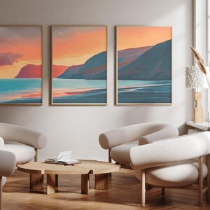 May include: Three framed landscape paintings of a beach scene with mountains in the background. The sky is a vibrant orange and pink, and the water is a turquoise blue. The paintings are hung on a white wall in a living room setting.