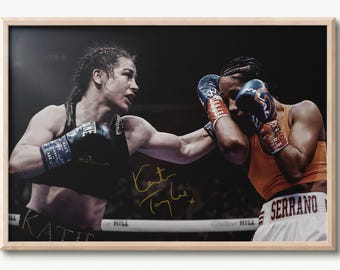 May include: A framed photograph of a boxing match featuring two athletes in a ring. One athlete wears a black top and black gloves, while the other wears an orange top, white shorts with the name SERRANO, and blue gloves. A gold signature is visible.
