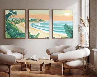 May include: A set of three framed wall art prints featuring a coastal landscape with sandy beaches, turquoise ocean waves, green palm leaves, and a distant sailboat under a soft peach-colored sky, displayed in a room with light-colored furniture.