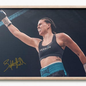 May include: Framed photograph of a boxer celebrating a win. The athlete wears a black sports bra with the "VERSION" logo, teal shorts, and boxing gloves. The image is signed in gold. The background is dark.