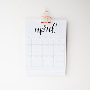 May include: A white calendar with the word "april" written in black cursive script. The calendar is hanging on a white wall with a gold clip.