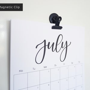 May include: A black magnetic clip holds a white calendar with the word "July" written in black cursive script. The calendar has the days of the week labeled at the top and numbers for the days of the month.