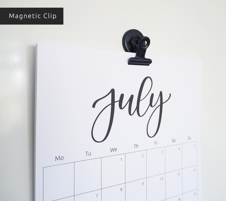 May include: A black magnetic clip holds a white calendar with the word "July" written in black cursive script. The calendar has the days of the week labeled at the top and numbers for the days of the month.