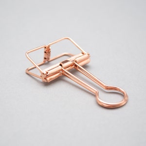 May include: A rose gold binder clip with a simple design. The clip is made of metal and has a smooth finish.