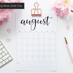May include: A white printable August calendar with a black handwritten title and a rose gold binder clip. The calendar is on a white surface with a keyboard, a pen, and pink flowers.