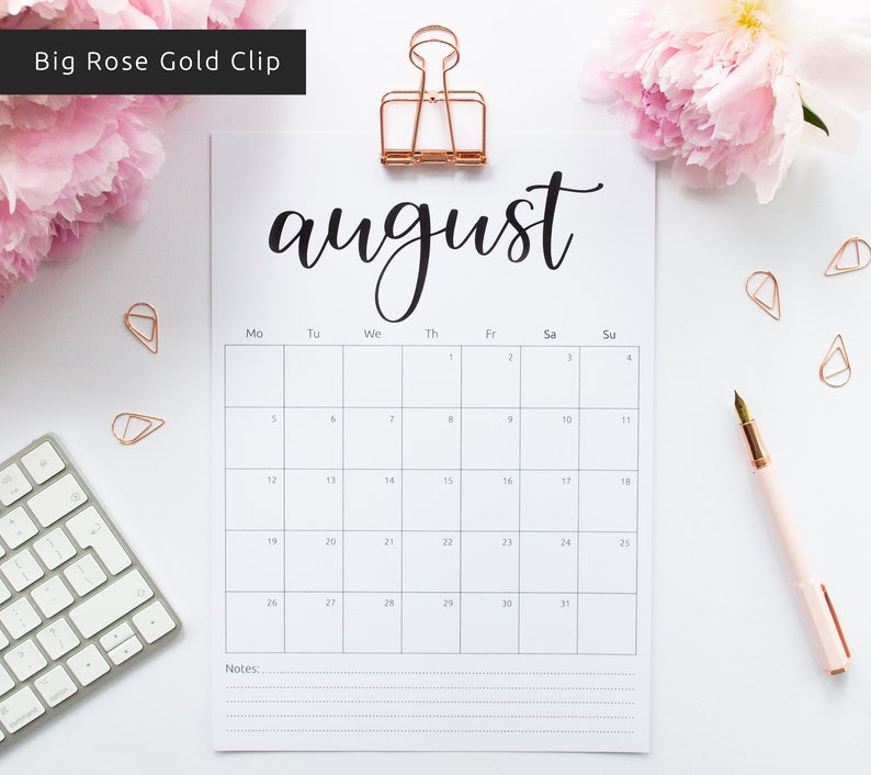 May include: A white printable August calendar with a black handwritten title and a rose gold binder clip. The calendar is on a white surface with a keyboard, a pen, and pink flowers.