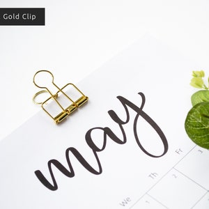 May include: A gold binder clip holding a calendar page with the word "may" written in black cursive script. The calendar page has the days of the week labeled and numbers for the days of the month.