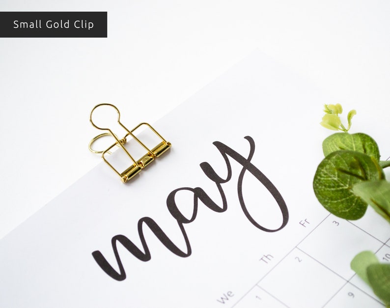 May include: A gold binder clip holding a calendar page with the word "may" written in black cursive script. The calendar page has the days of the week labeled and numbers for the days of the month.