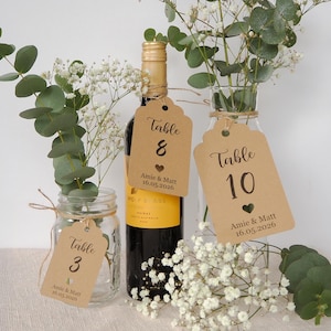 May include: A wedding table setting featuring kraft paper table number tags. The tags are attached to a wine bottle and glass vases with floral arrangements. The tags read "Table 3", "Table 8", and "Table 10".