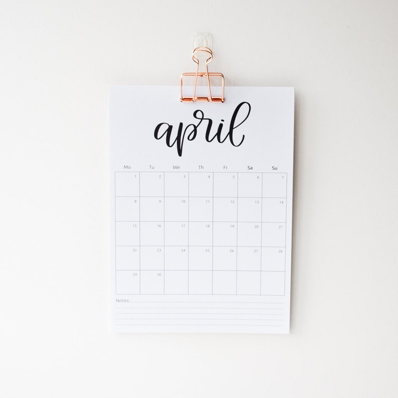 May include: A white calendar with the word "april" written in black cursive script. The calendar is hanging on a white wall with a gold clip.