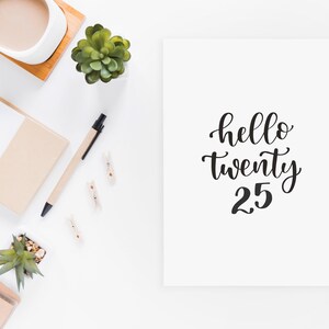 May include: White card with black calligraphy that reads "hello twenty 25". The card is on a white surface with a potted succulent, a cup of coffee, a notepad, a pen, and wooden clothespins.