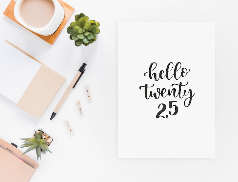 May include: White card with black calligraphy that reads "hello twenty 25". The card is on a white surface with a potted succulent, a cup of coffee, a notepad, a pen, and wooden clothespins.