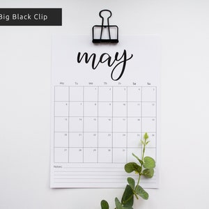 May include: A black and white calendar with the word "may" written in cursive at the top. The calendar is hanging from a black binder clip. The calendar has the days of the week labeled at the top and the numbers 1 through 31 listed in the grid below. There is a small green plant in the bottom right corner of the image.