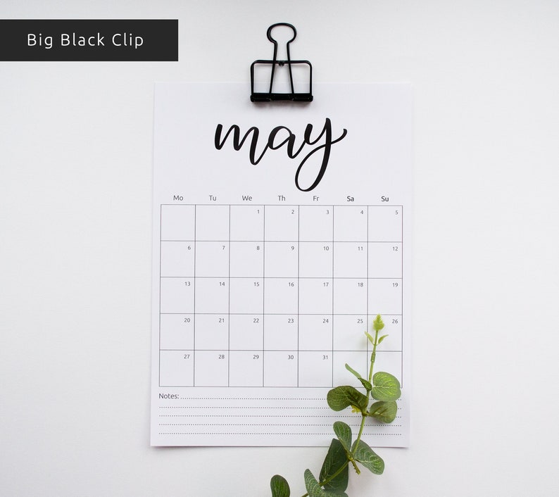 May include: A black and white calendar with the word "may" written in cursive at the top. The calendar is hanging from a black binder clip. The calendar has the days of the week labeled at the top and the numbers 1 through 31 listed in the grid below. There is a small green plant in the bottom right corner of the image.